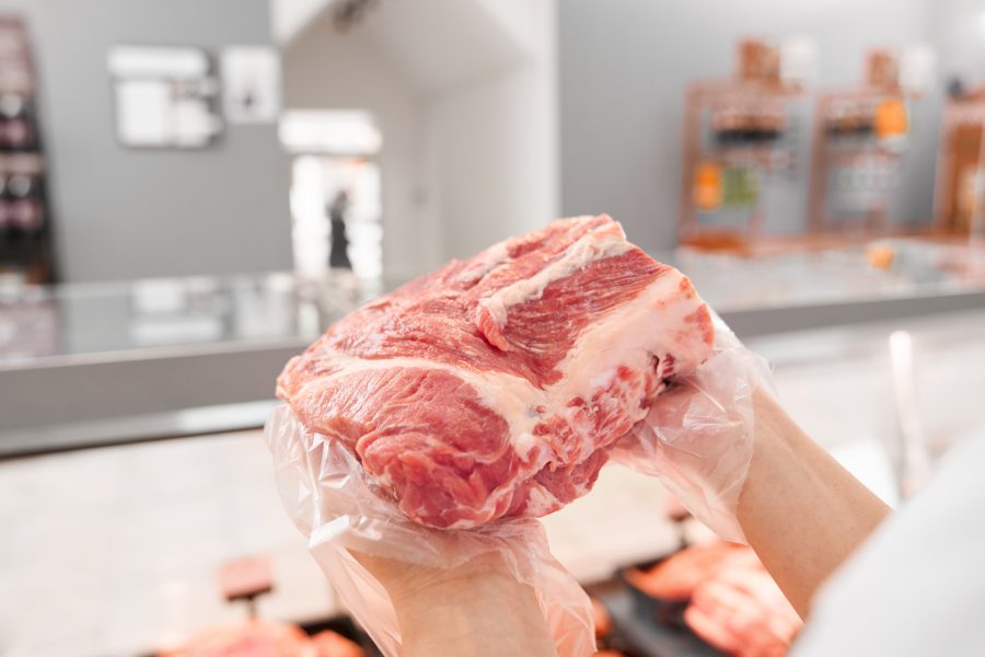 Foto close-up tangan menggunakan sarung tangan plastik transparan memegang potongan besar daging sapi mentah segar di sebuah toko daging dengan latar belakang yang buram