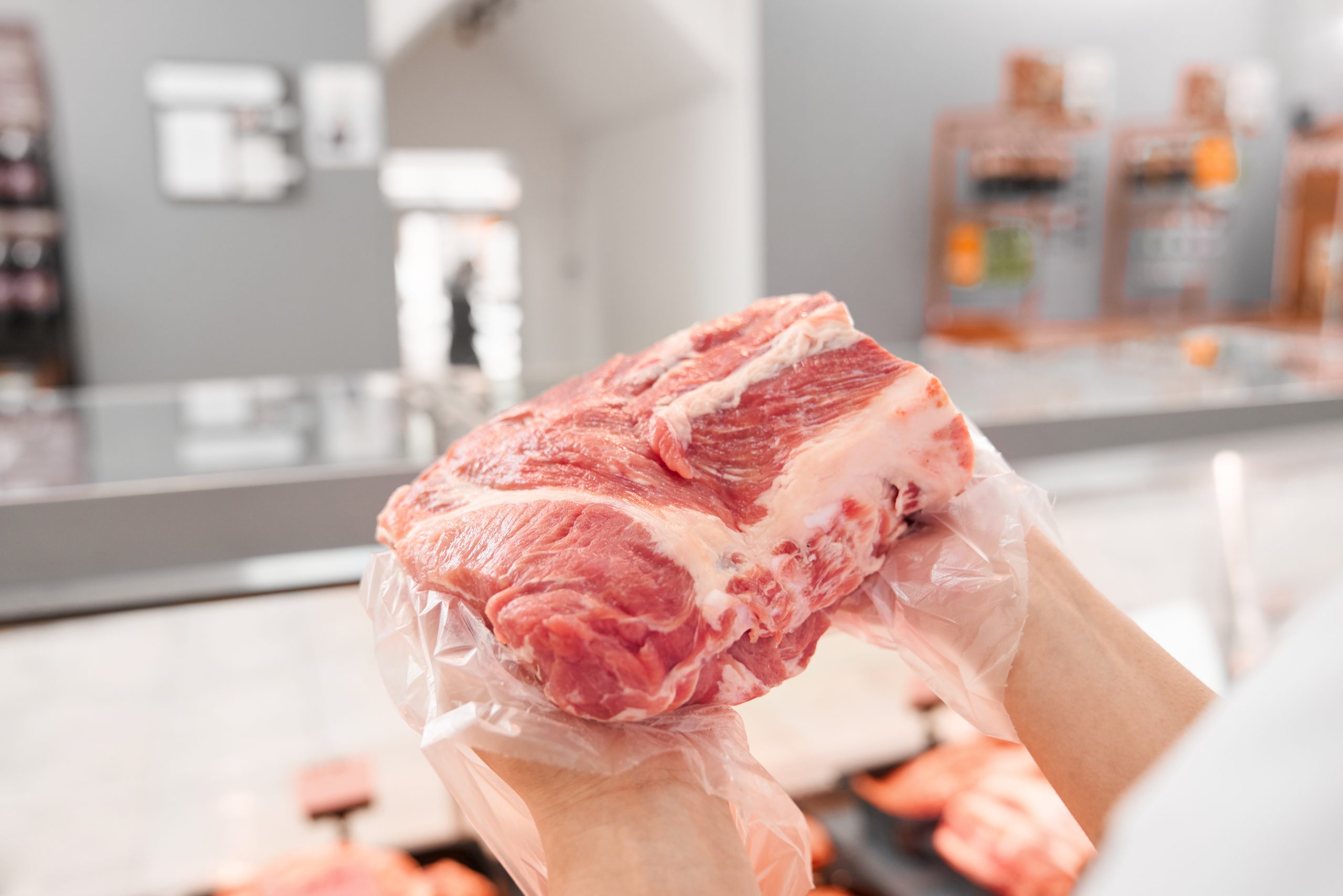 Foto close-up tangan menggunakan sarung tangan plastik transparan memegang potongan besar daging sapi mentah segar di sebuah toko daging dengan latar belakang yang buram